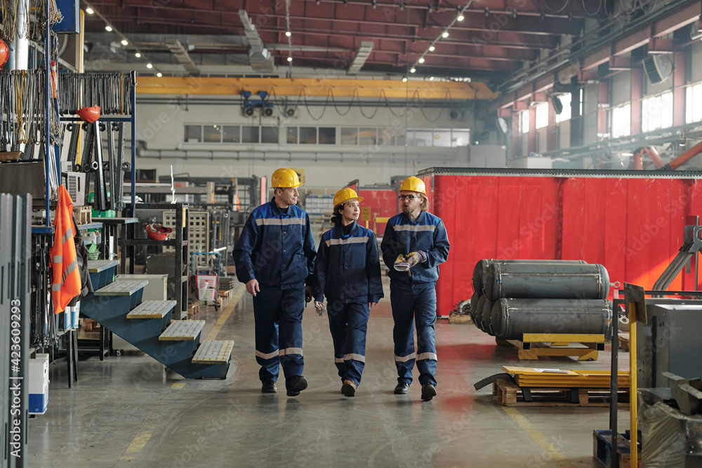 Group of workers or engineers of contemporary factory moving along large workshop with industrial equipment and having conversation