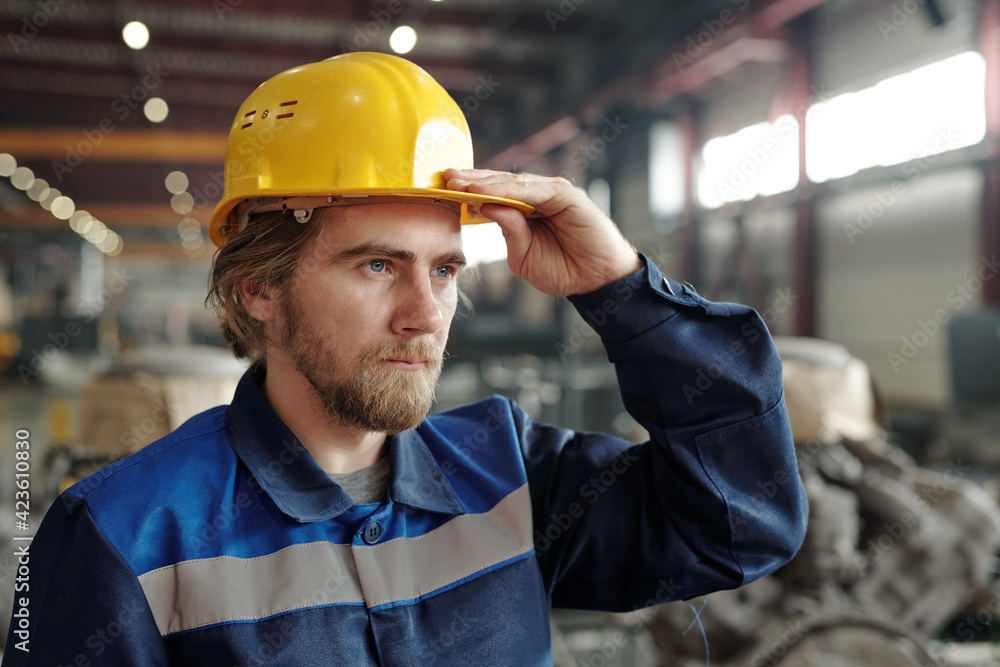 Young bearded male engineer or factory worker in protective helmet standing in front of camera while working in industrial plant Young bearded male engineer or factory worker in protective helmet standing in front of camera while working in industrial plant