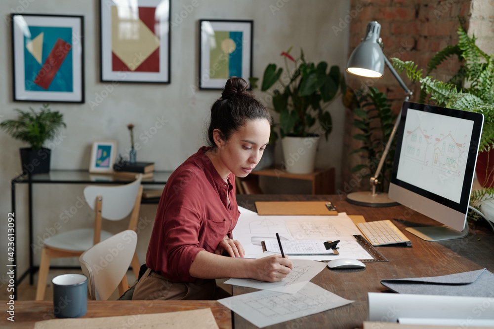 Young serious female architect with pencil drawing sketch on paper while sitting by table in front of computer screen with picture of house Young serious female architect with pencil drawing sketch on paper while sitting by table in front of computer screen with picture of house
