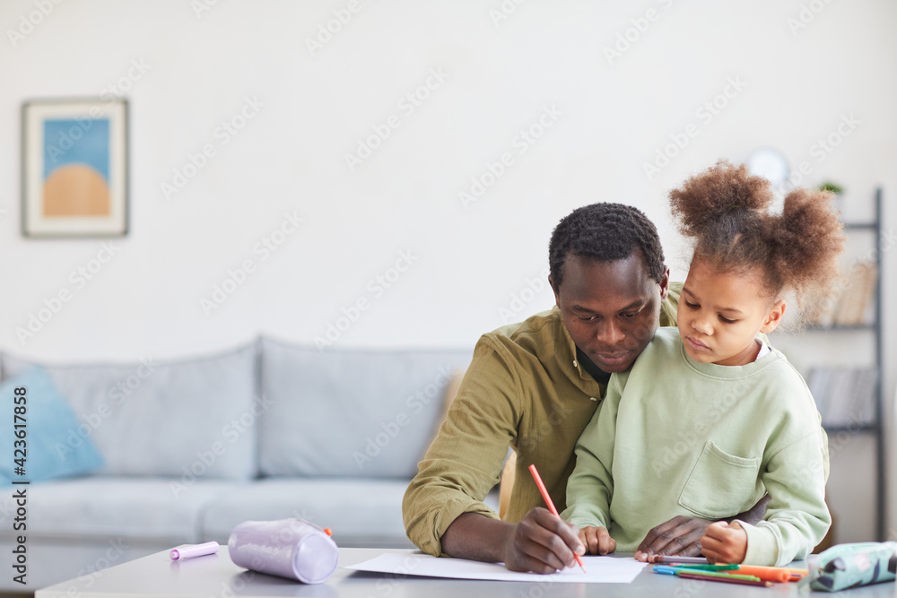 Front view portrait of caring African-American father helping cute girl doing howework or drawing at desk in minimal home interior, copy space Front view portrait of caring African-American father helping cute girl doing howework or drawing at desk in minimal home interior, copy space