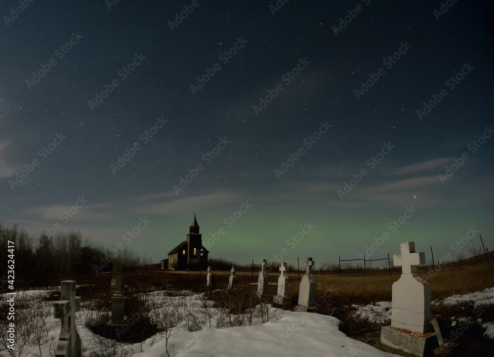 Bleak looking dilapidated graveyard with an old church in the background under a starry night.  Scattered clouds and weak Aurora can be seen in the sky.
