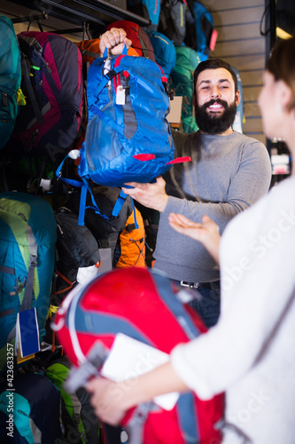 Smiling guy displays a woman a big rucksacks while shopping for sporting goods