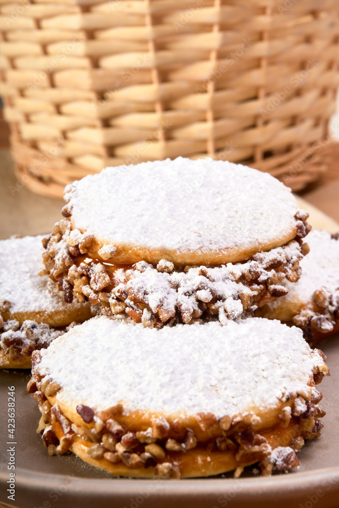 argentinian alfajores with dulce de leche and nuts, sprinkled with icing sugar on a brown plate. on one side a cup of coffee on a rustic wooden table. a traditional argentinian dessert.