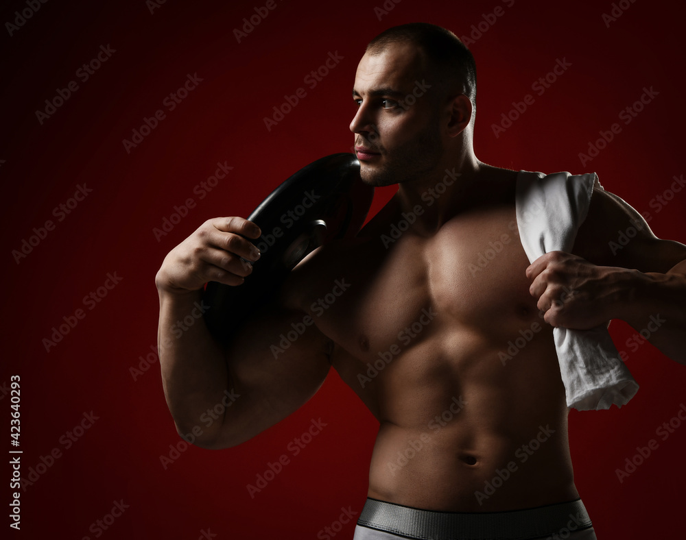 Portrait of muscular man, bodybuilder standing shirtless with naked chest holding towel and weight for barbell on shoulder and looks aside over red background