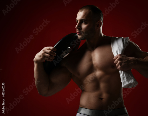 Portrait of muscular man, bodybuilder standing shirtless with naked chest holding towel and weight for barbell on shoulder and looks aside over red background © Dmitry Lobanov