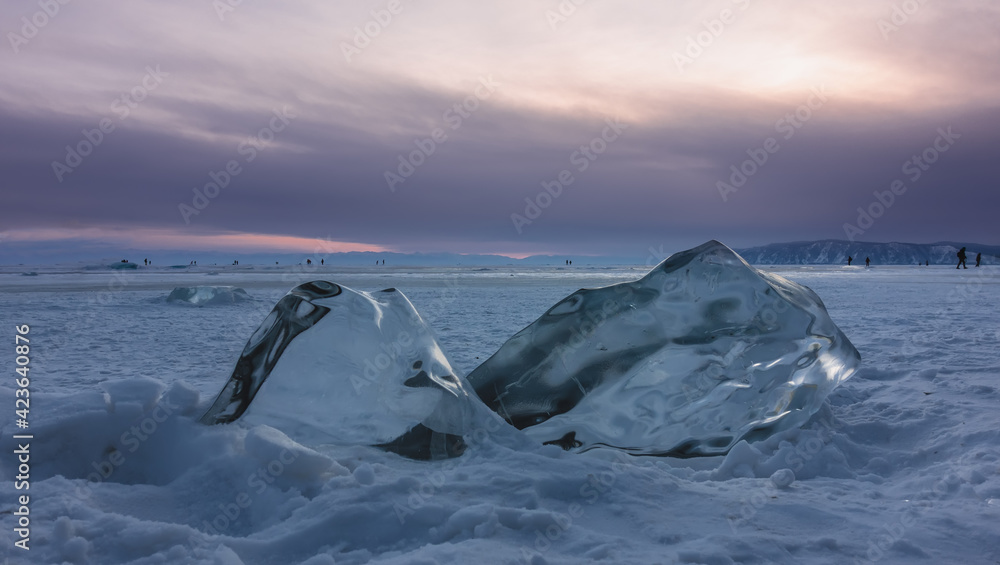 Fototapeta Duże przezroczyste lśniące kępy lodu na zamarzniętym i zaśnieżonym jeziorze. Zbliżenie. W oddali widać ludzi chodzących po lodzie. Zachód słońca różowe niebo. Kontury gór. Bajkał