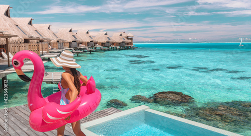 Travel vacation fun tourist woman enjoying luxury summer holidays at Bora Bora overwater bungalow swimming with flamingo pool toy float at infinity pool by turquoise ocean. Tahiti getaway destination.