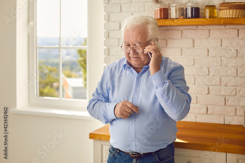 White-haired senior man making phone call and ordering food or meal delivery standing in the kitchen. Mature grandfather staying home and keeping in touch with family talking with them on mobile photo