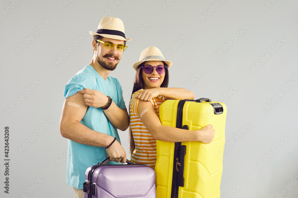 Young people ready for safe summer holiday journey. Studio portrait of happy couple holding travel cases, smiling and showing arms after receiving COVID-19 vaccine. Coronavirus vaccination concept - obrazy, fototapety, plakaty 