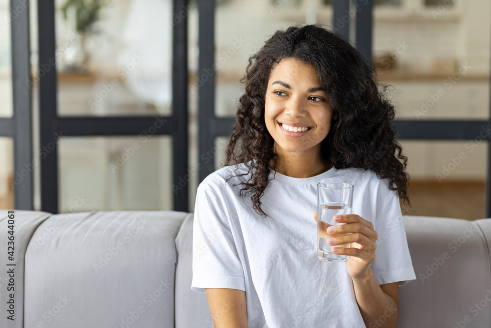 Portrait pretty young african american woman smiling sit on couch holding glass of still or mineral water, healthy lifestyle concept Portrait pretty young african american woman smiling sit on couch holding glass of still or mineral water, healthy lifestyle concept