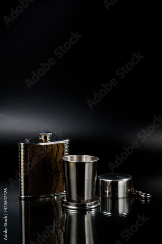 A metal pocket flask for alcoholic beverages with a folding metal cup, shot against a dark background.