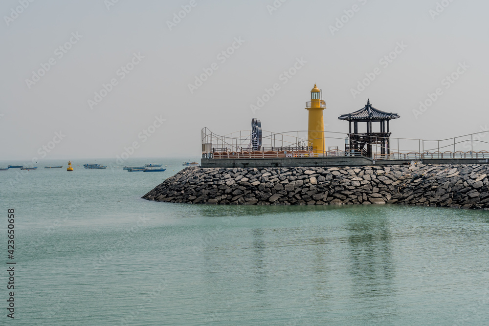 Yellow lighthouse beside oriental gazebo on seaside pier with gray sky in background.