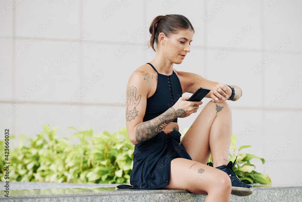 Fit pretty young woman sitting outdoors and synchronizing fitness trekker and smartphone to control her activity Fit pretty young woman sitting outdoors and synchronizing fitness trekker and smartphone to control her activity
