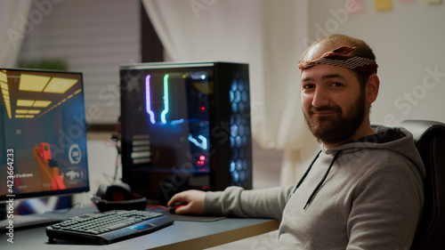 Portrait of happy man looking at camera smiling playing online FPS shooter videogame for virtual tournament. Cyber performing on powerful computer with RGB keyword during championship at home