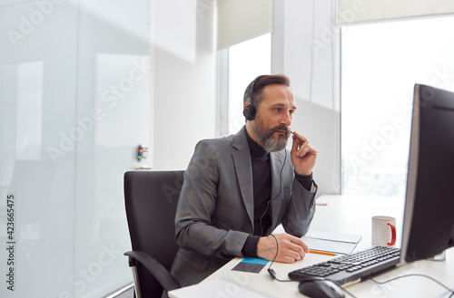 Happy matured man with headphones is sitting in a light white office