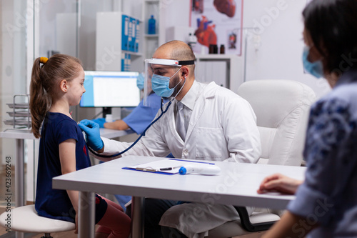 Child looking at specialist doctor while listening heart with stethoscope for medical examination. Pediatrician doctor with protection gloves and mask against covid19 talking about treatment disease