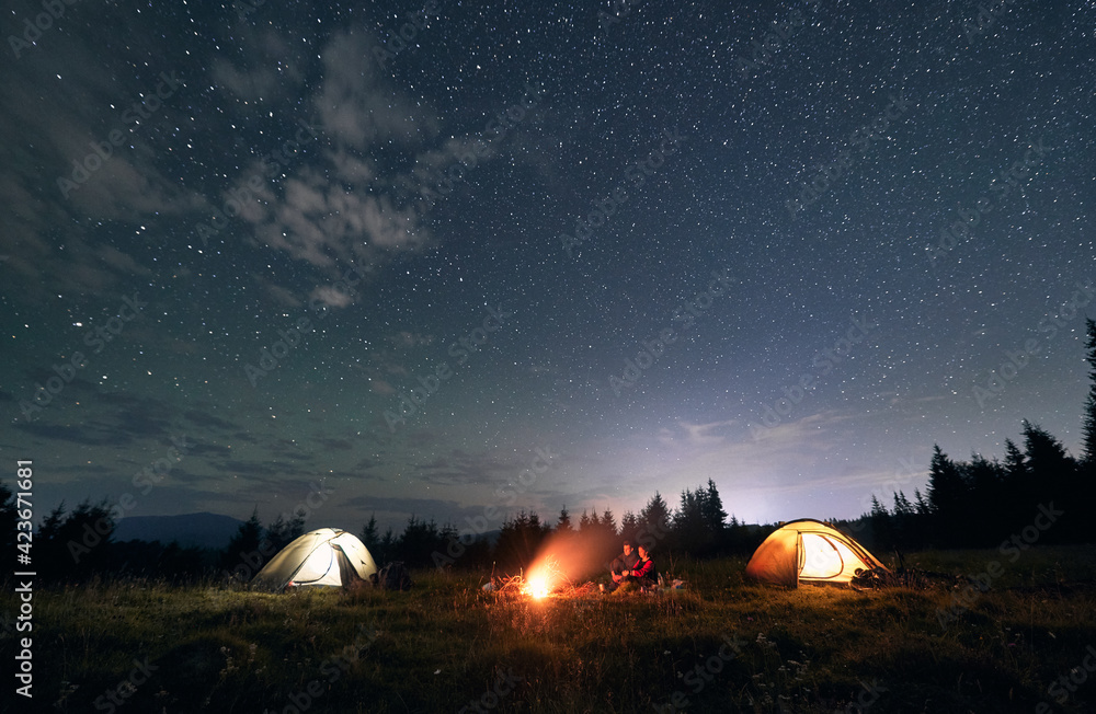Hikers standing near campfire under beautiful starry sky.