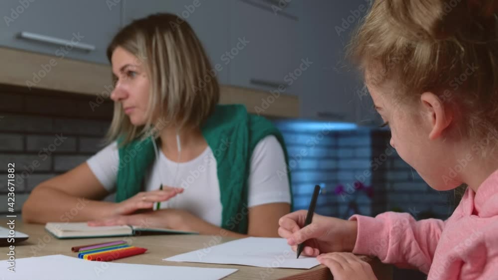 Caucasian daughter is doing her homework while her mother is sitting near and working on the laptop Caucasian daughter is doing her homework while her mother is sitting near and working on the laptop