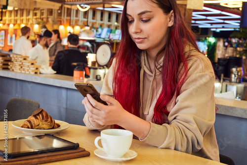 Beautiful woman with red hair is typing a message in the phone while sitting in a restaurant over a cup of coffee with a croissant.