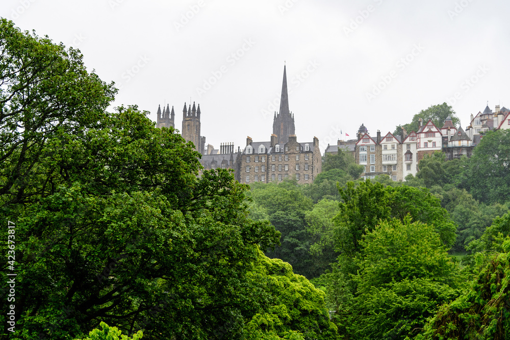 Row of old historic buildings behind large green trees in Edinburgh, Scotland in a cloudy rainy summer day.