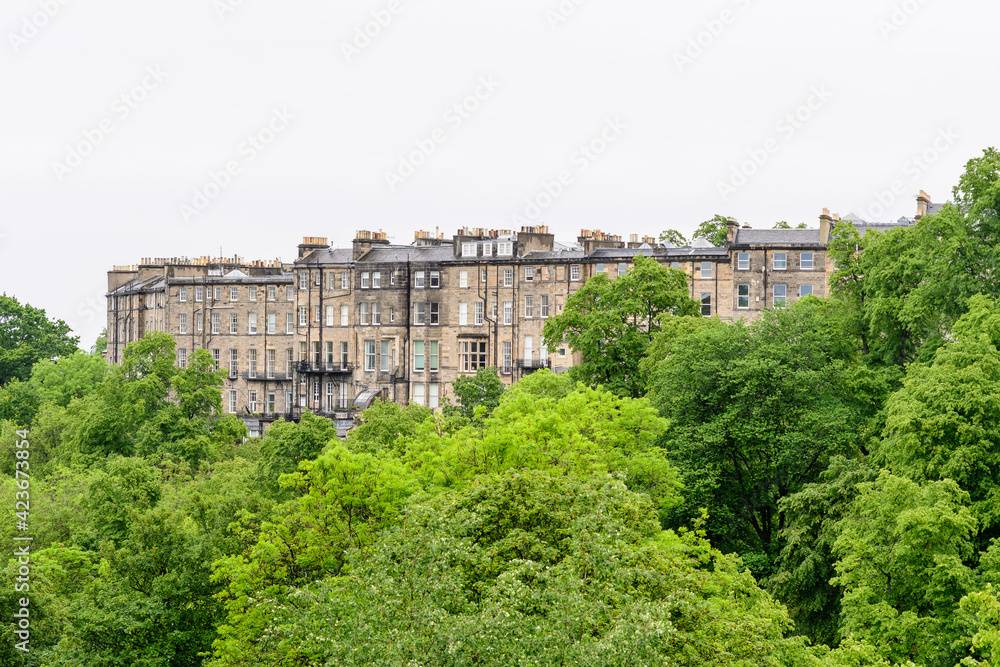 Row of old historic buildings behind large green trees in Edinburgh, Scotland in a cloudy rainy summer day.
