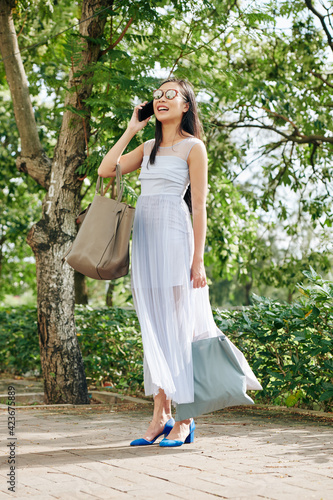 Happy joyful young Asian woman in sunglsses standing in city park with textile bags and talking on phone with friend or family member