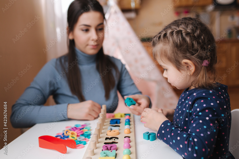 Fototapeta premium Little girl play in educational game for children with mom. Wooden game with different colors and numbers. Intecactive game for clever kids. Mom spend time with daughter during qurantine while