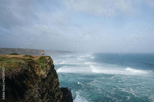 Small unrecognizable figure of woman standing on a cliff at ocean on windy day