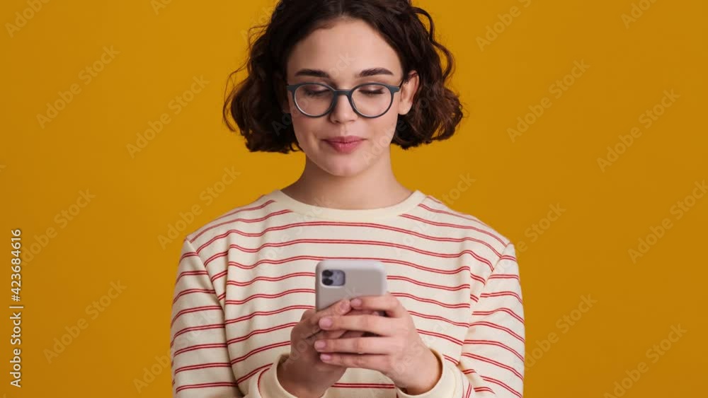 A pretty nice young teenager girl in eyeglasses is using her smartphone standing isolated over yellow background in studio A pretty nice young teenager girl in eyeglasses is using her smartphone standing isolated over yellow background in studio