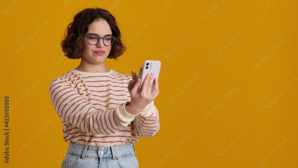 A pretty nice young teenager girl in eyeglasses is taking selfie-photo using her smartphone standing in the side isolated over yellow background in studio A pretty nice young teenager girl in eyeglasses is taking selfie-photo using her smartphone standing in the side isolated over yellow background in studio