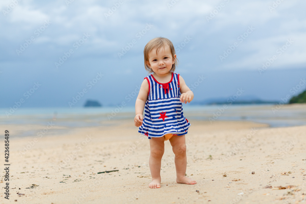 Toddler baby doing her first steps on sand near the bank. Little cute girl playing on the beach. Small child enjoying vacation by the sea or ocean. Travelling with kids concept. Photo with copy space Toddler baby doing her first steps on sand near the bank. Little cute girl playing on the beach. Small child enjoying vacation by the sea or ocean. Travelling with kids concept. Photo with copy space