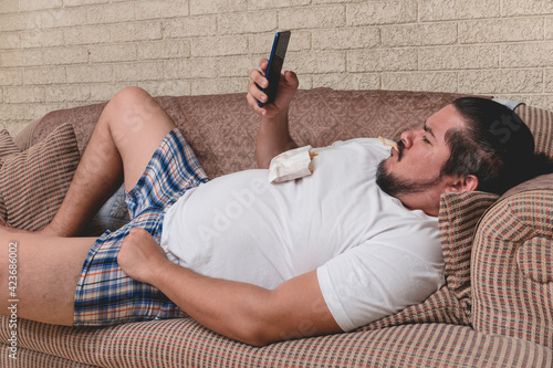 A lazy and overweight bum munches on some french fries while watching a video or browsing social media on the phone. Couch potato on the sofa. photo