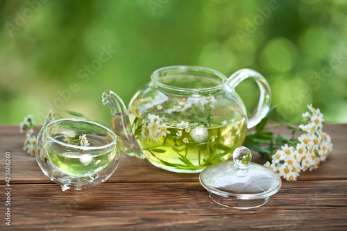 Herbal tea with achillea salicifolia flowers in a glass teapot and glass thermo cup on a green bokeh background.