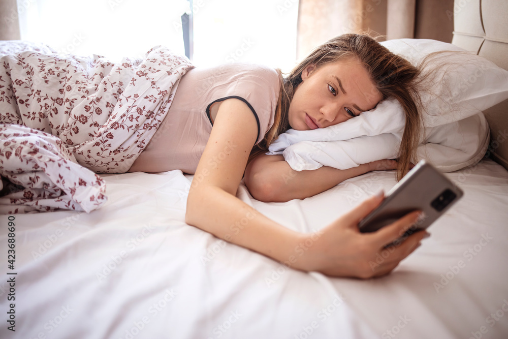 Woman lying in bed reaching to turn off the alarm on her mobile phone in morning. Sleepy woman being woken up by smartphone alarm. Young woman waking up in bed and checking her smartphone Woman lying in bed reaching to turn off the alarm on her mobile phone in morning. Sleepy woman being woken up by smartphone alarm. Young woman waking up in bed and checking her smartphone