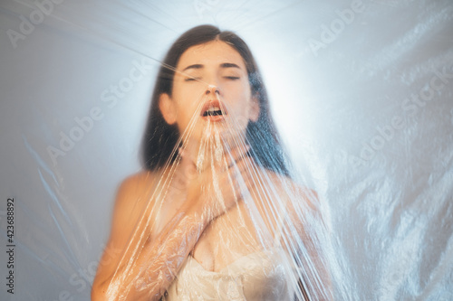 Shortness of breath. Defocused portrait. Air pollution. Allergy asthma. Suffering woman with gasp touching throat behind transparent wrinkled polyethylene film texture on light background out of focus