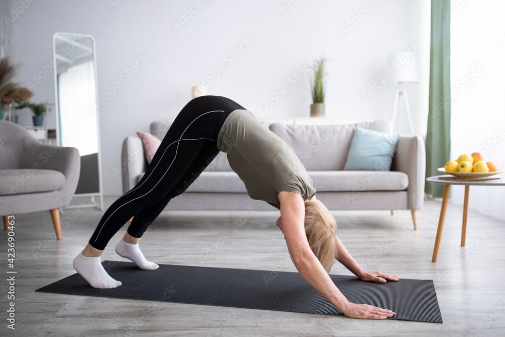 Domestic yoga practice. Flexible mature lady standing in downward facing dog asana, exercising on sports mat at home Domestic yoga practice. Flexible mature lady standing in downward facing dog asana, exercising on sports mat at home