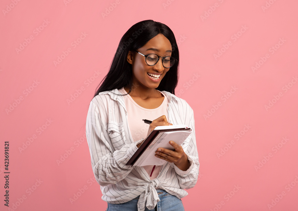 Fototapeta premium Portrait of smart black lady in glasses writing in notebook on pink studio background