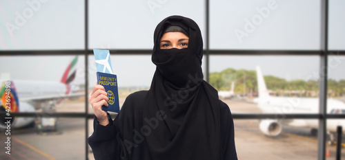 safe travel, tourism and people concept - muslim woman in hijab with air ticket and immunity passport over airport background