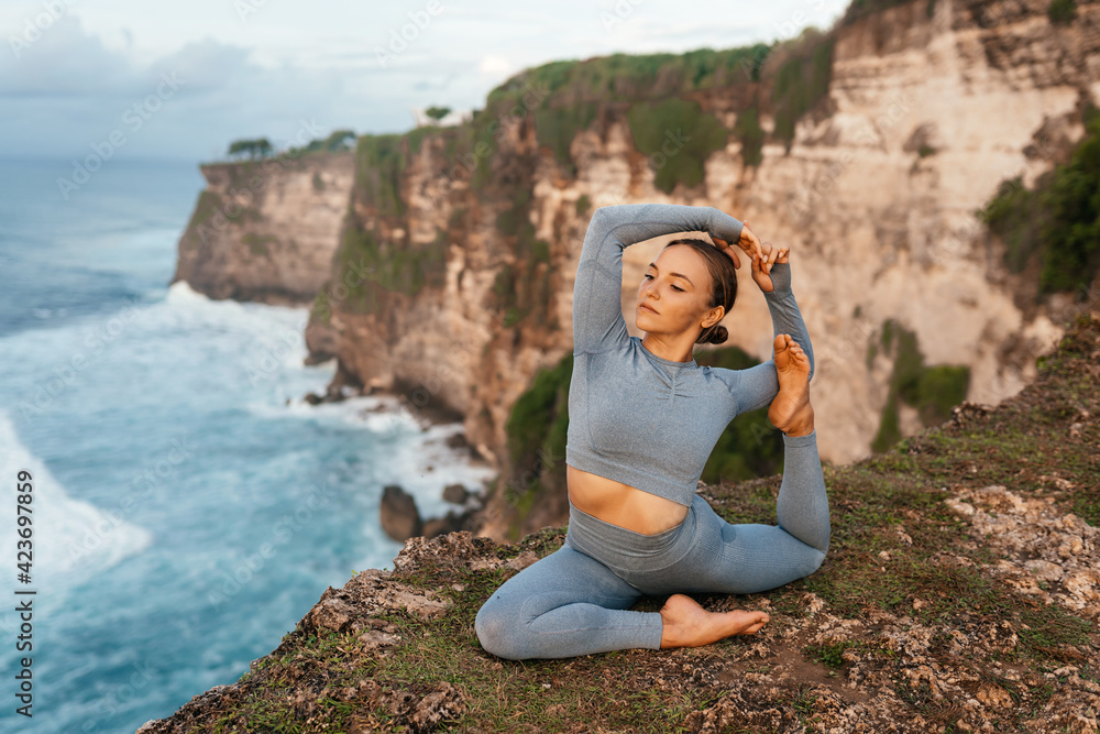 Beautiful woman doing yoga on a cliff, behind an amazing view in the ocean