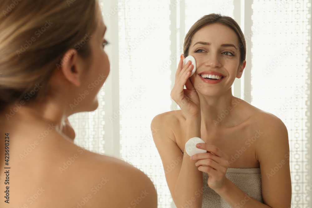 Young woman with cotton pads cleaning her face near mirror in bathroom Young woman with cotton pads cleaning her face near mirror in bathroom