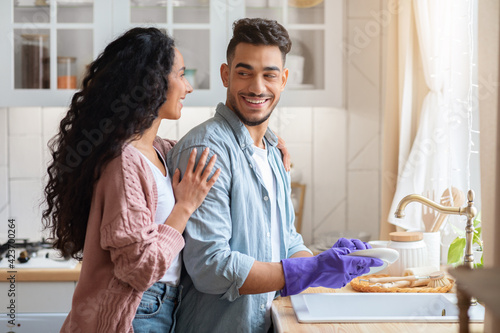 Fotografie Loving Arab Wife Embracing Her Husband While He Washing Dishes In Kitchen
