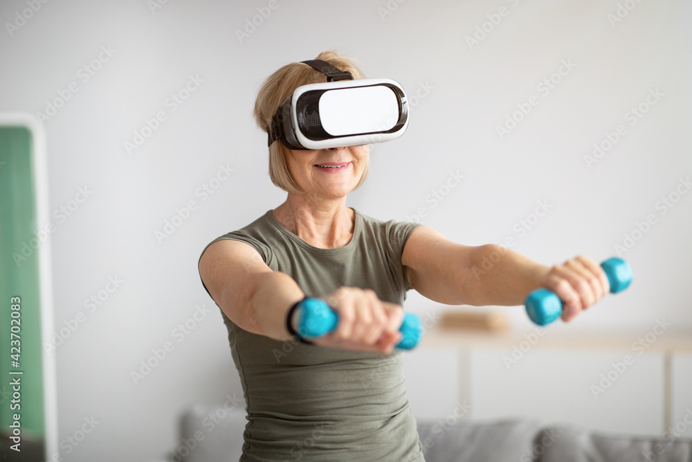 Virtual workout concept. Fit senior lady in VR glasses doing exercises with dumbbells at home Virtual workout concept. Fit senior lady in VR glasses doing exercises with dumbbells at home