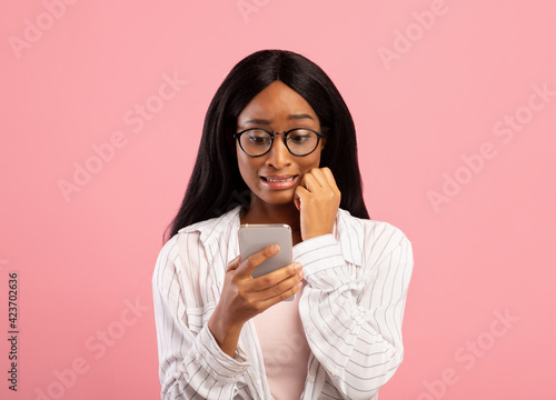Oops. Embarassed black woman looking at mobile phone screen in confusion on pink studio background photo