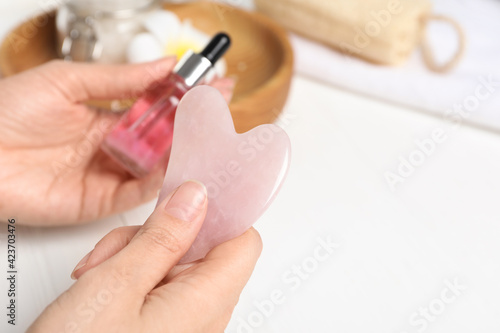 Woman holding gua sha tool and cosmetic at white wooden table  closeup. Space for text