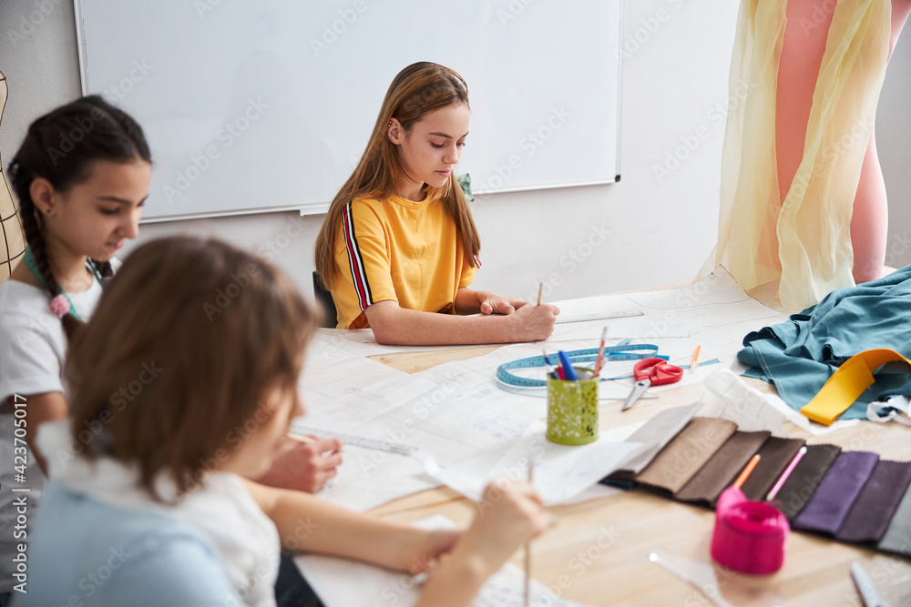Fototapeta premium Adorable girls drawing clothes patterns in sewing workshop