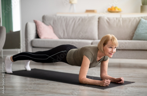 Domestic workout concept. Athletic senior woman standing in elbow plank on yoga mat at home