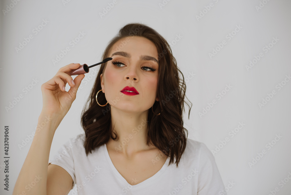 Woman with makeup and red lips applying black mascara on eyelashes with brush. Photos of appealing brunette girl on white background Woman with makeup and red lips applying black mascara on eyelashes with brush. Photos of appealing brunette girl on white background