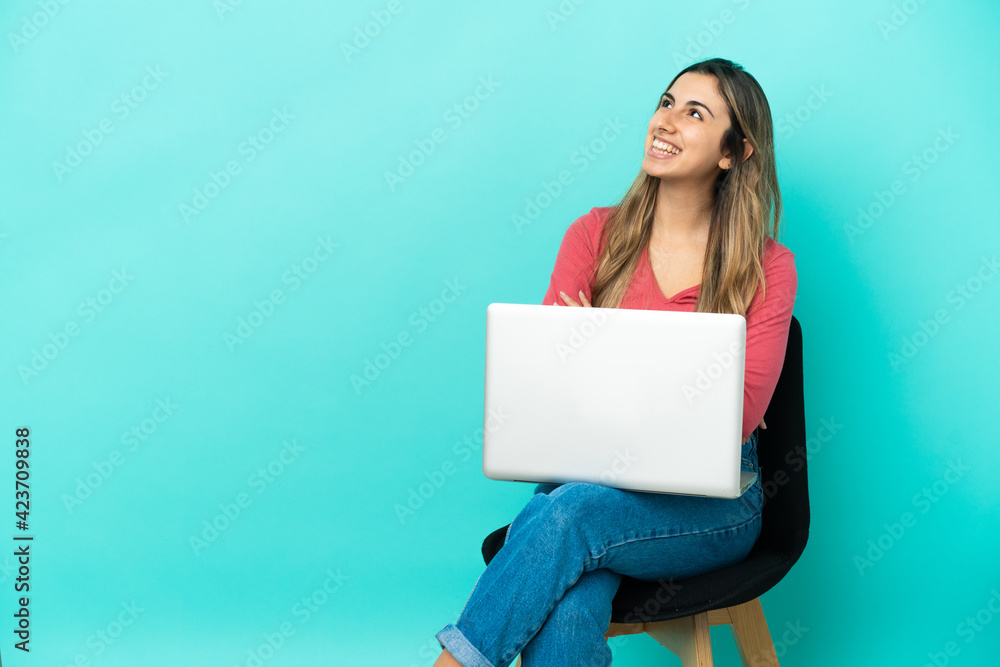 Young caucasian woman sitting on a chair with her pc isolated on blue background looking up while smiling Young caucasian woman sitting on a chair with her pc isolated on blue background looking up while smiling