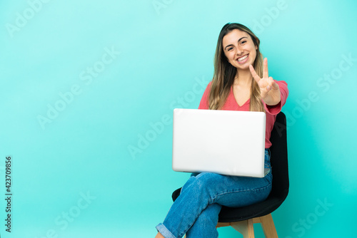 Young caucasian woman sitting on a chair with her pc isolated on blue background smiling and showing victory sign © luismolinero