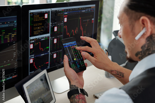 Trader checking stock market charts on computer screens and buying stocks via application on smartphone